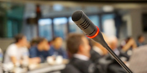 Beschlüsse öffentlicher Sitzungen: Mikrofon / Im Hintergrund kann man unscharf Menschen bei einer Sitzung erklennen.